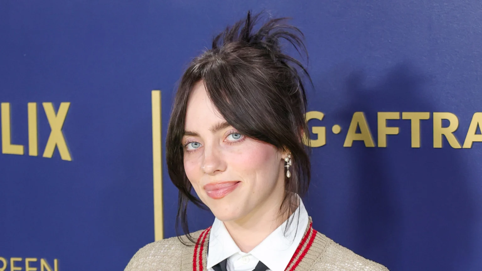 Billie Eilish smiles faintly, wearing a white shirt, black tie, and a tan cardigan with red stripes, against a blue wall with gold letters for Netflix and SAG-AFTRA.