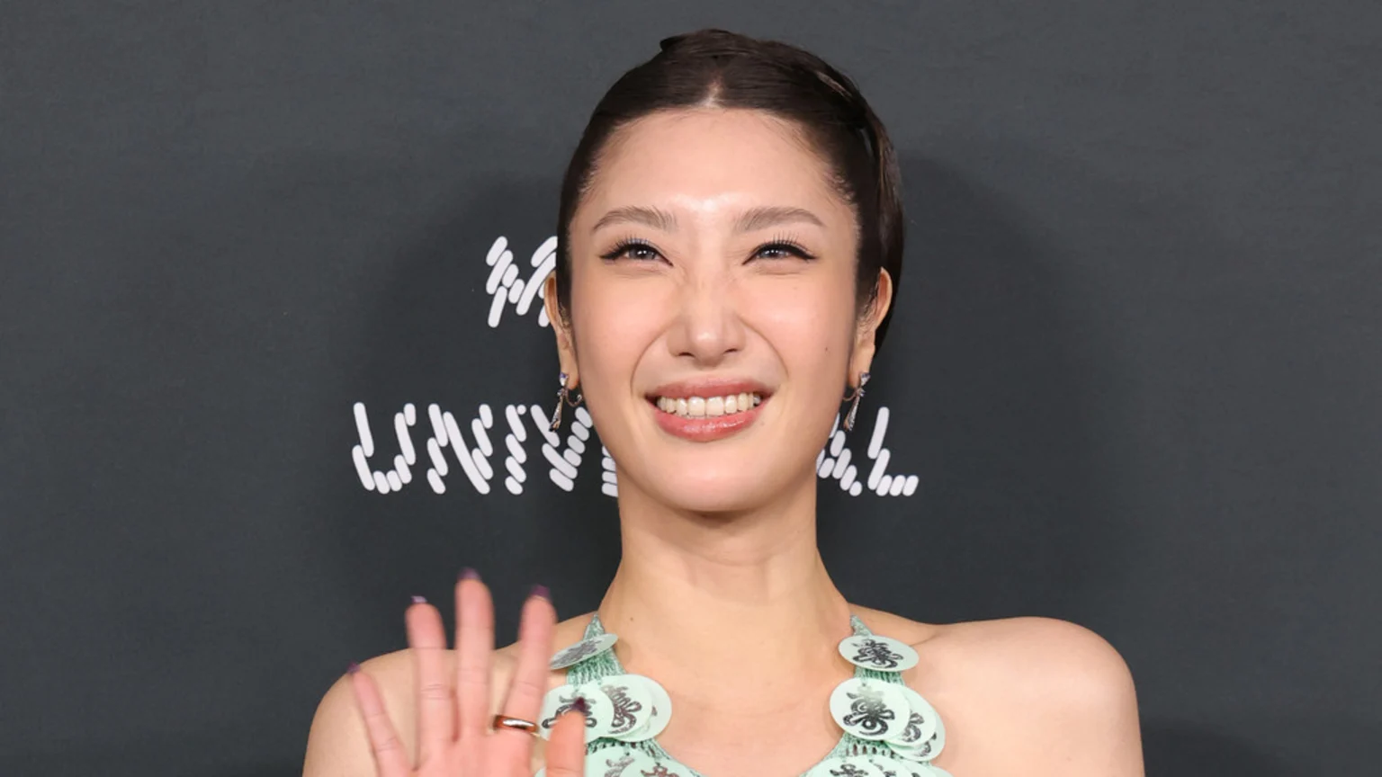 A smiling woman with dark hair pulled back, prominent eyeliner, and a light green top waves at the camera against a dark background with stylized white text.