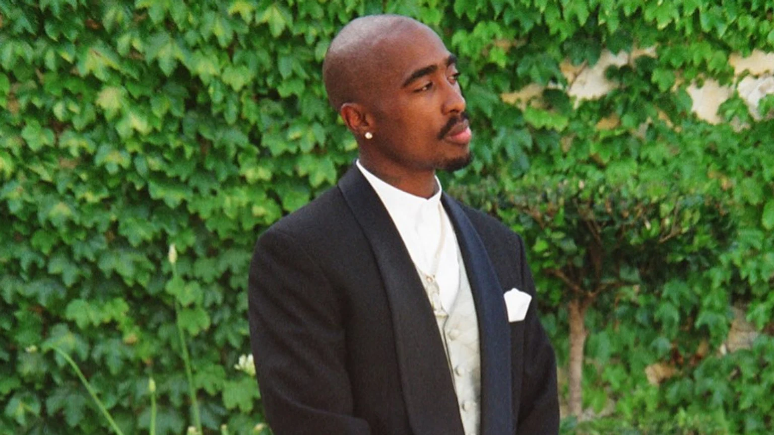 A formal headshot of Tupac Shakur standing outdoors in a garden or courtyard, dressed in a black tuxedo jacket, white shirt, and light waistcoat, with a silver chain and pearl earring, against a lush ivy-covered wall and small shrub background.