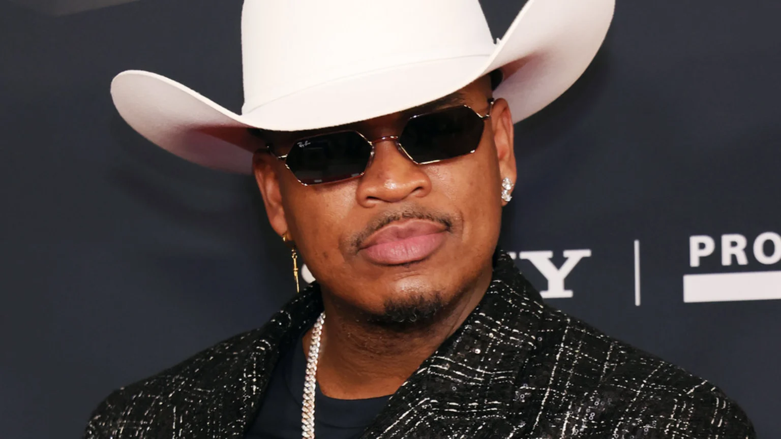 A man identified as Ne-Yo, wearing a white cowboy hat, Ray-Ban sunglasses, a mustache, goatee, and a dark textured jacket with a silver chain, looks forward.