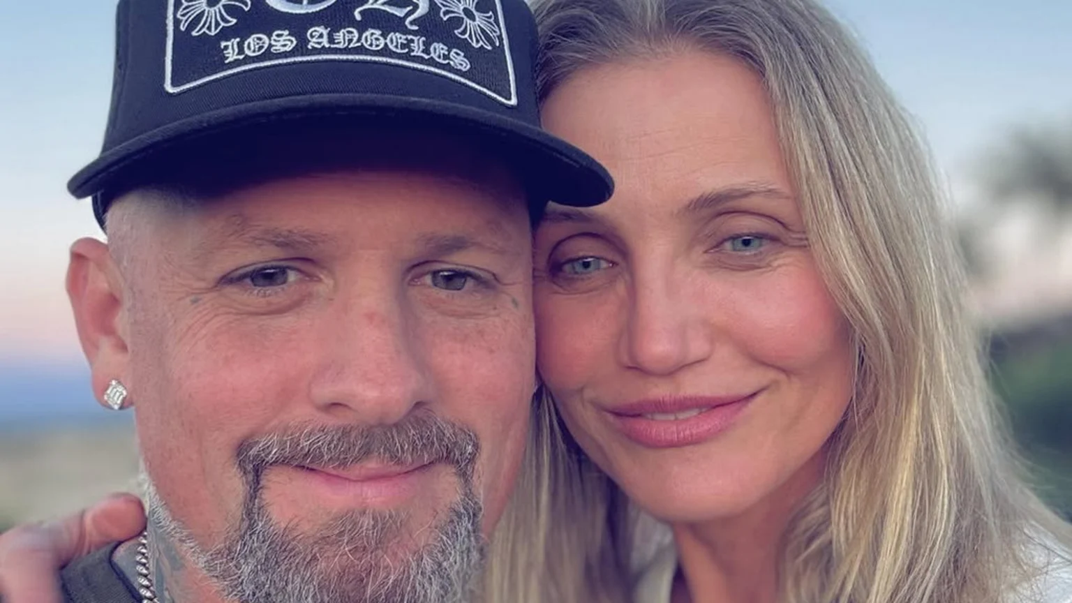 A close-up selfie shows Benji Madden, wearing a black "LOS ANGELES" cap and beard, leaning against Cameron Diaz, who has blonde hair and a faint smile, with a soft outdoor background.