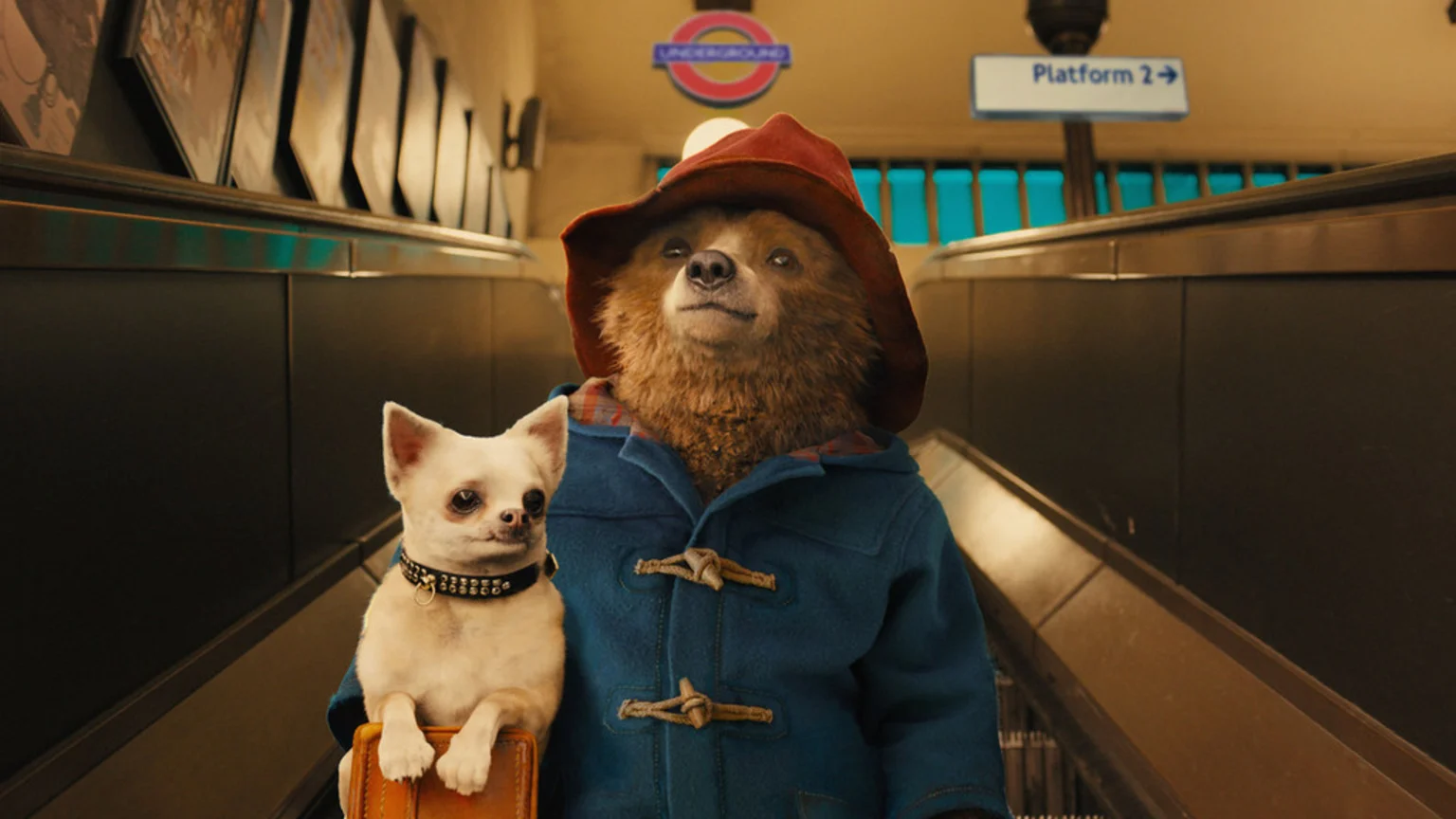 Paddington Bear in a red hat and blue coat stands on an escalator with a small white dog sitting on a suitcase, with London Underground signs in the background.