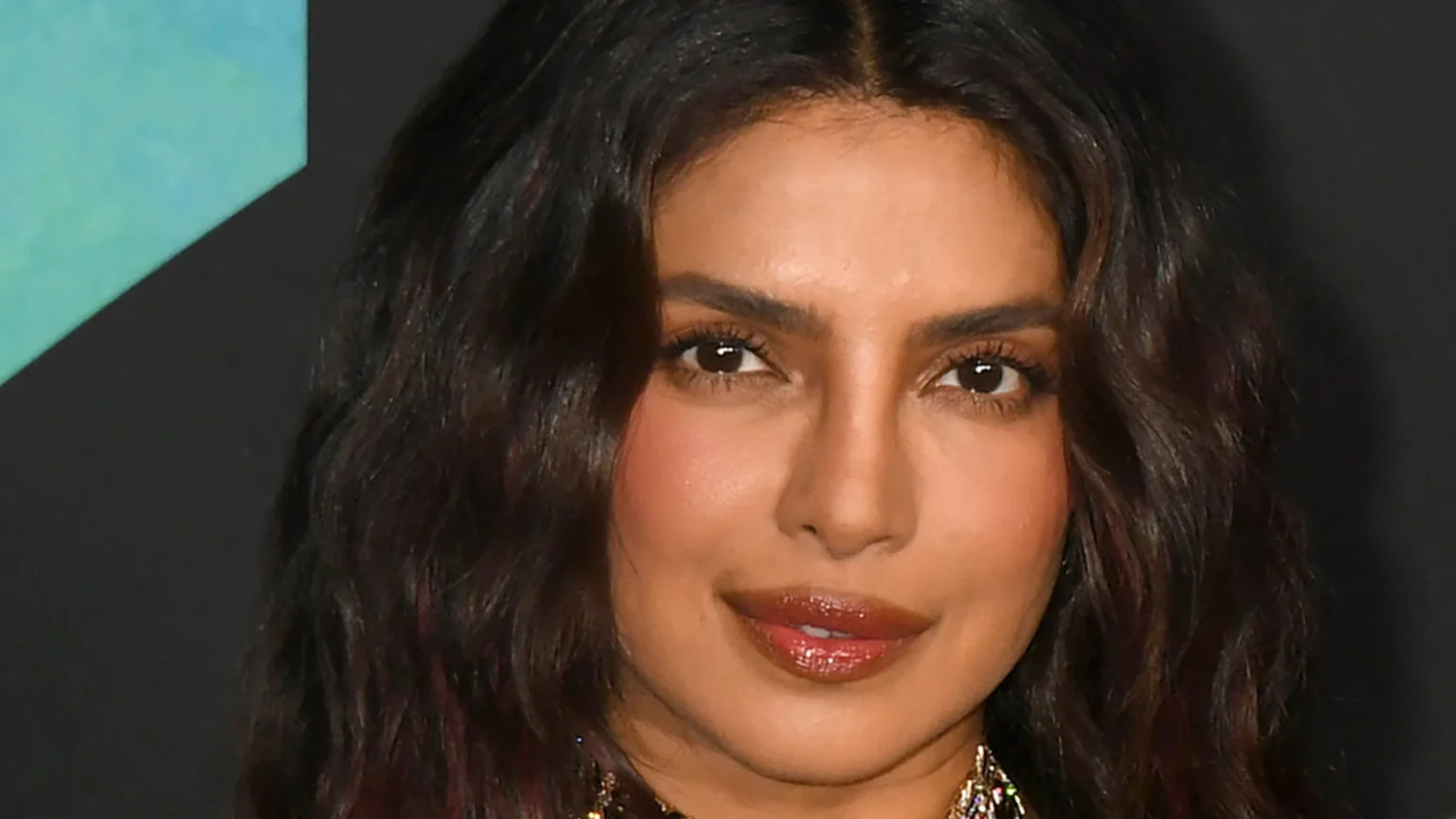 Close-up of Priyanka Chopra smiling slightly, with dark wavy hair, warm skin, dark eyes, and glossy lips, against a dark background with a hint of blue-green on the left.