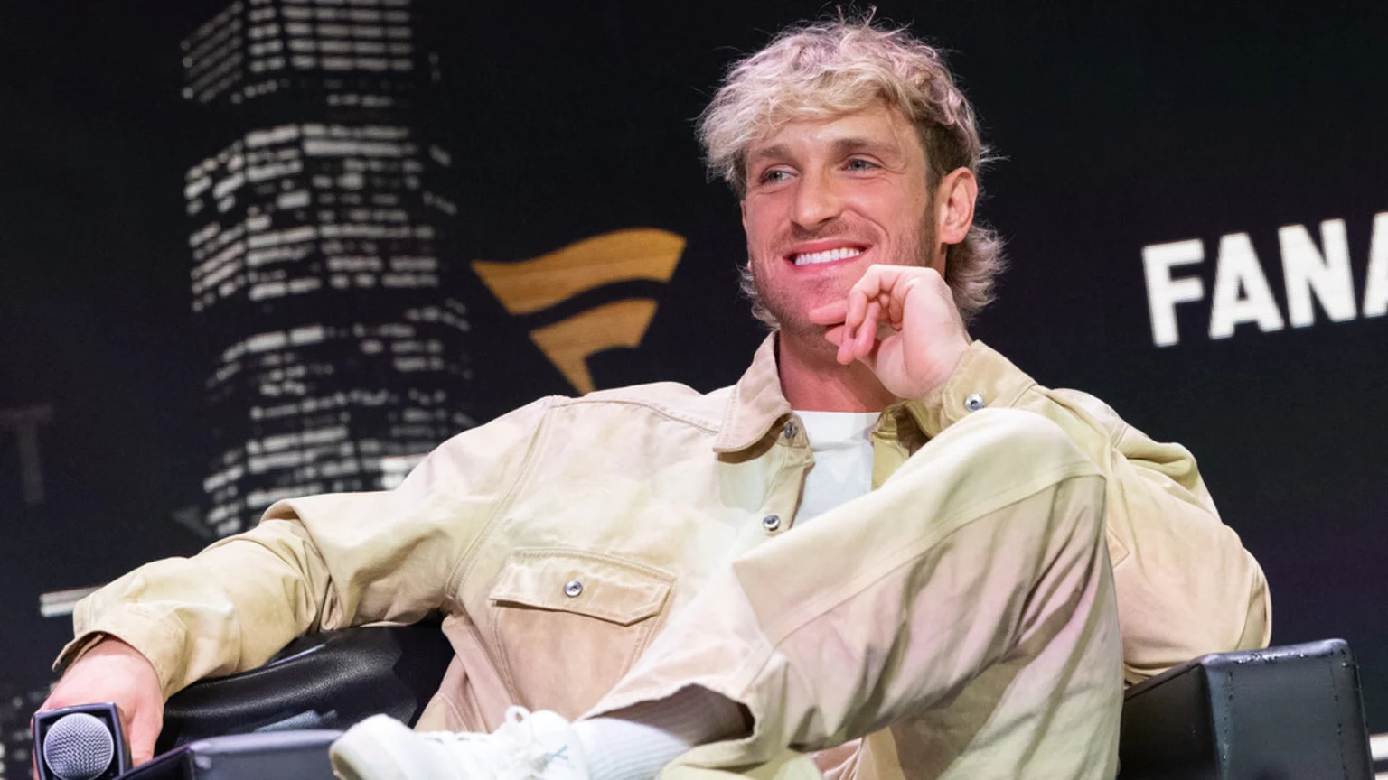 Logan Paul, wearing light tan jacket and pants with white athletic shoes, sits in a black leather armchair holding a silver microphone. The dim backdrop features white skyscraper outlines and a golden Fanatics logo, with bright focused lighting on him.