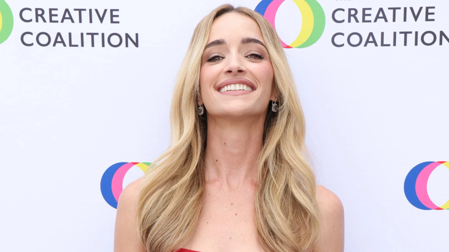 A smiling Brianne Howey, with long blonde hair and earrings, poses in front of a white banner displaying "THE CREATIVE COALITION" logo.