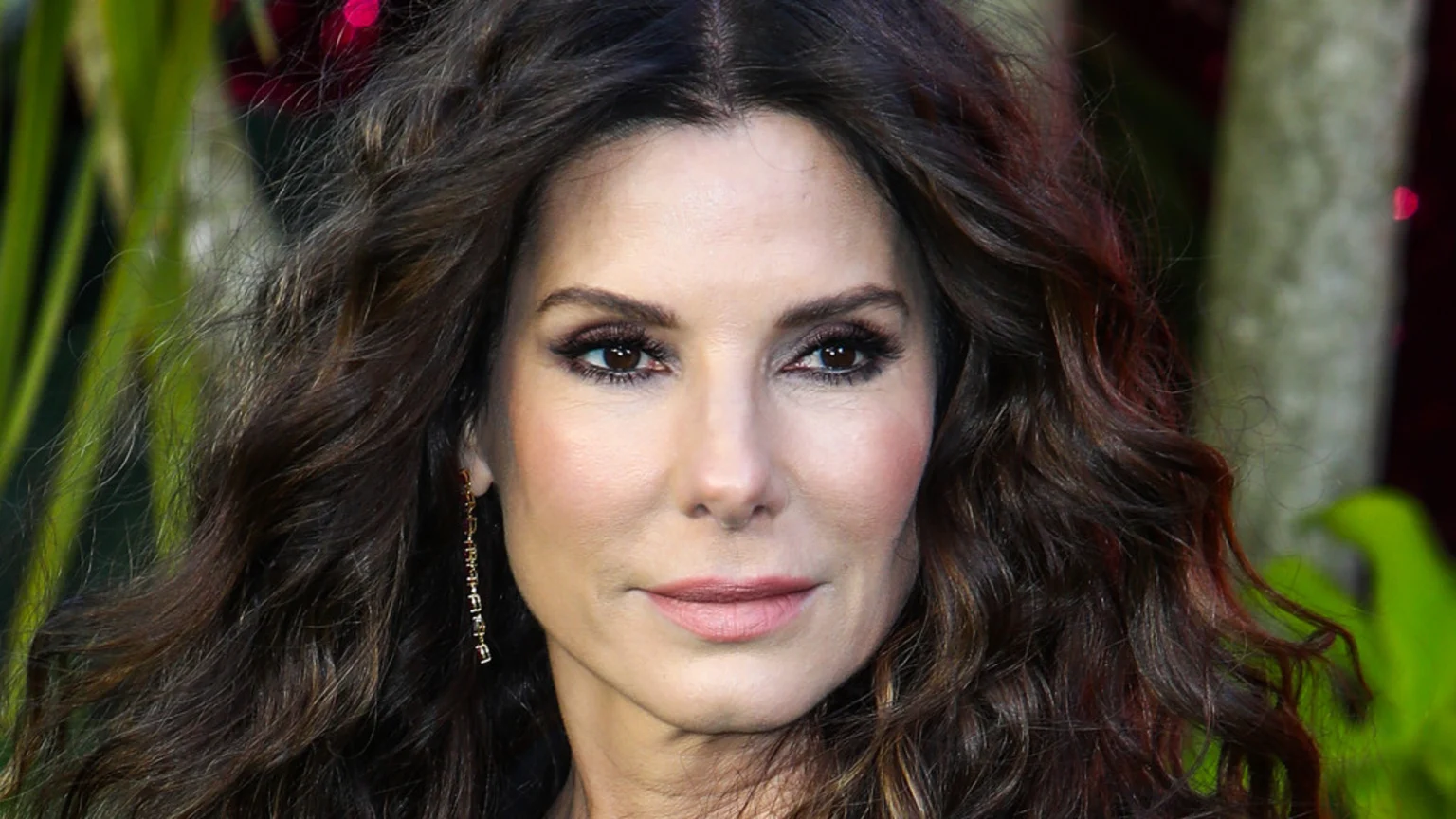 Close-up of actress Sandra Bullock, facing forward with a neutral expression, her long dark wavy hair parted down the middle, wearing makeup, and an earring on her left ear.