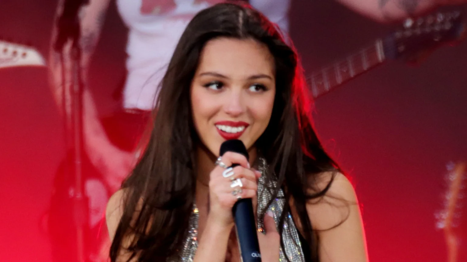 Olivia Rodrigo, with long dark hair and red lipstick, smiles while holding a microphone and wearing rings, including one spelling "FUT", against a blurred red background.