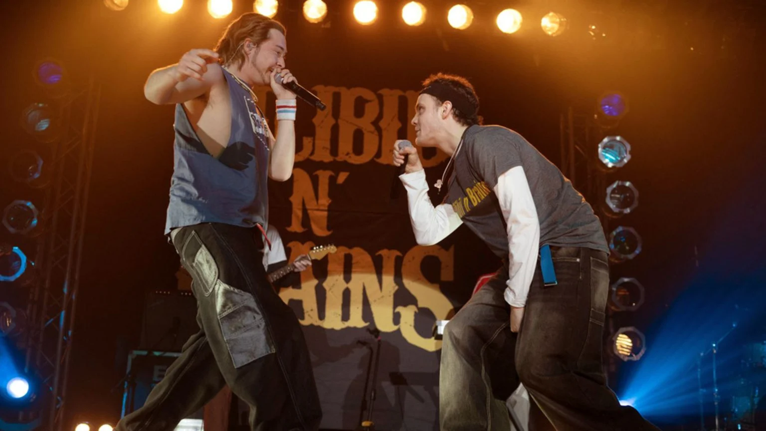 Two men on a stage perform with microphones, illuminated by stage lights and backed by a banner.