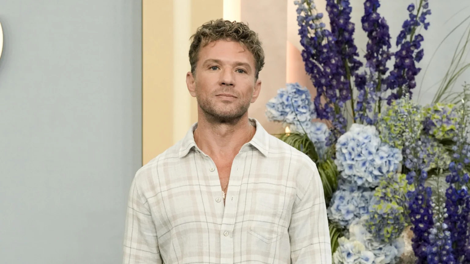 A man with curly brown hair and stubble wears a light plaid shirt, looking directly at the camera, with blue and purple floral arrangements in the background.
