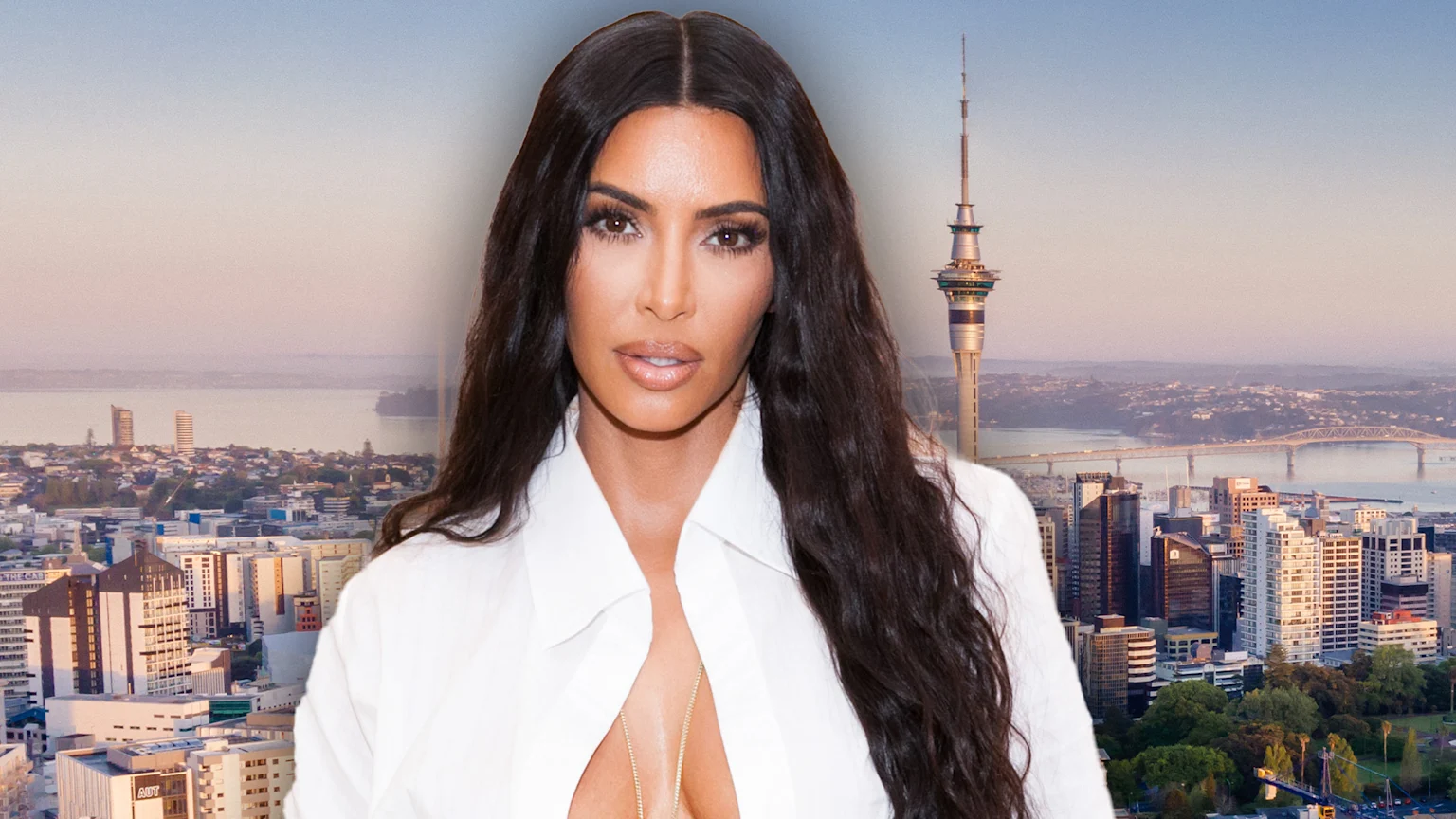Kim Kardashian, wearing a white shirt and gold necklace, is overlaid on an aerial view of Auckland, New Zealand, featuring the Sky Tower and Harbour Bridge.