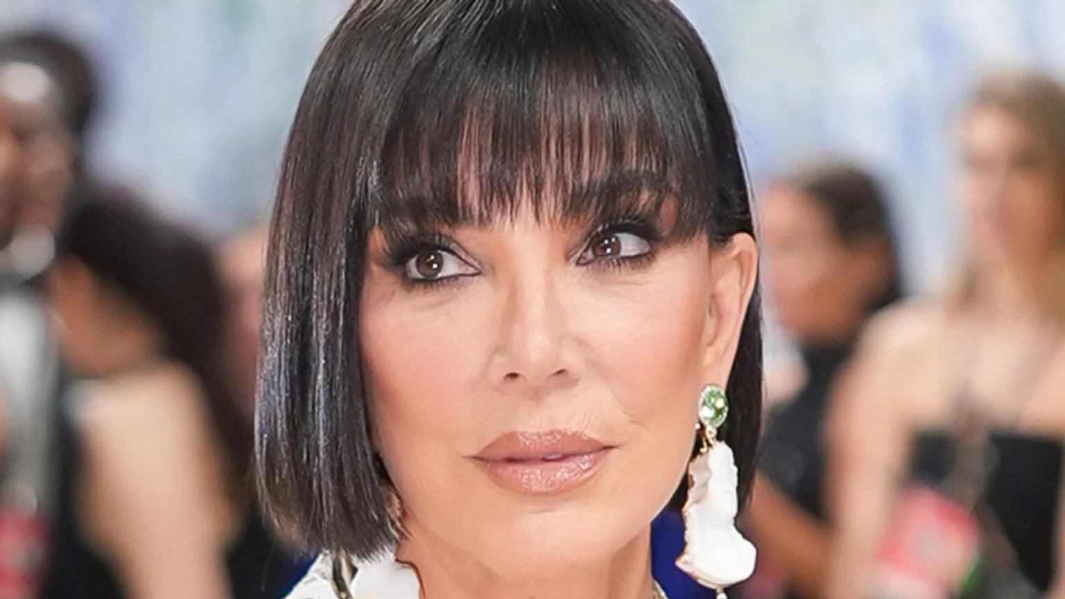 A close-up of Kris Jenner, a woman with short dark hair and bangs, striking brown eyes, glossy nude lips, and a large green and white ornate dangling earring.