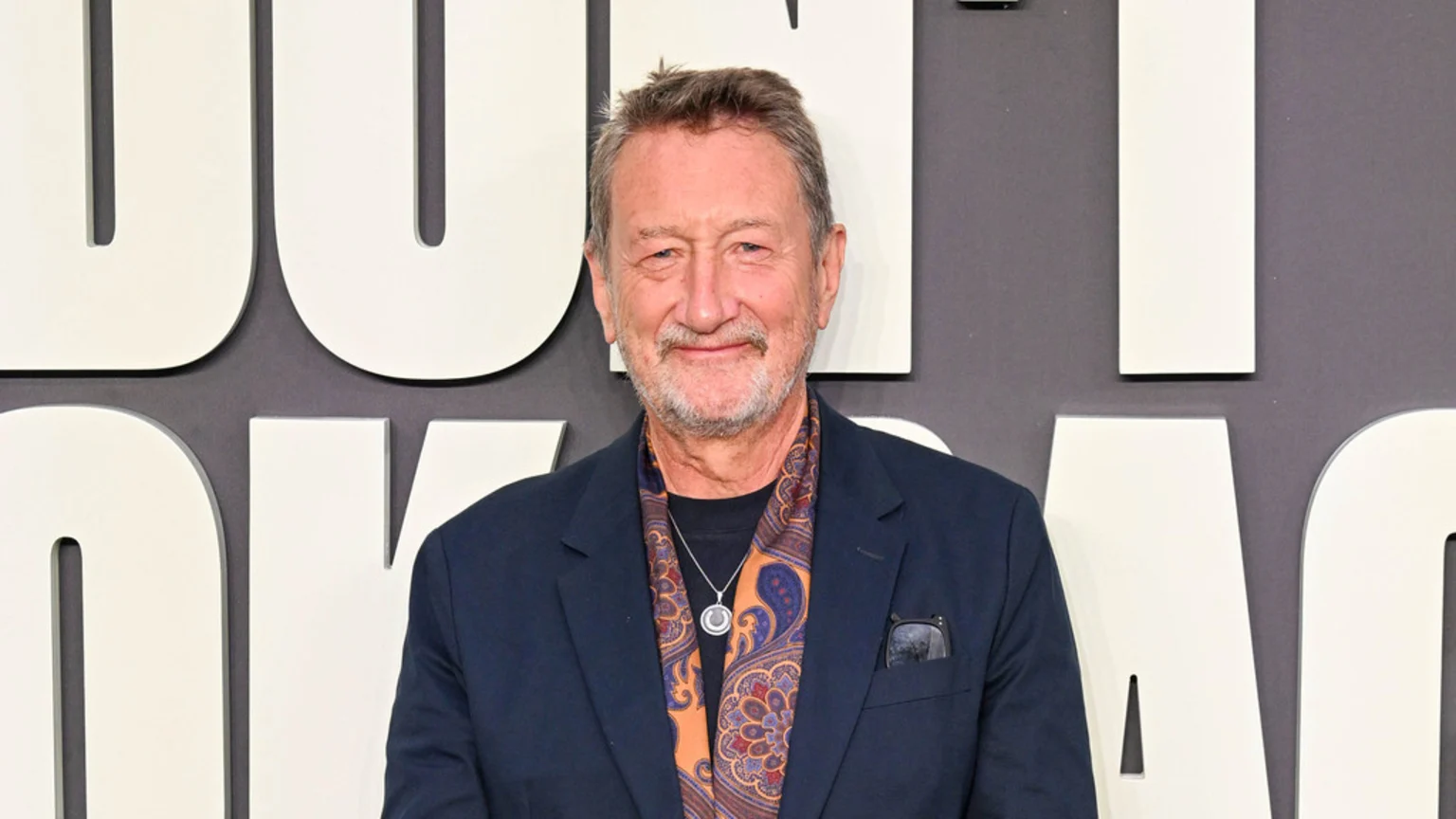 A smiling man with a beard, wearing a dark blue suit jacket, a paisley scarf, and a silver necklace, stands in front of large white letters on a grey background.