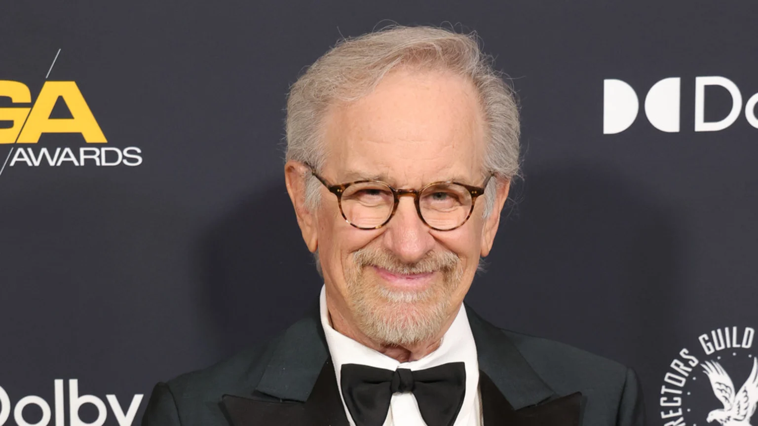 A close-up portrait of Steven Spielberg smiling warmly at an indoor Directors Guild Awards event. He wears tortoise-shell rimmed glasses, a dark suit with black satin lapel, and a black bow tie. The background features logos for GA Awards, Dolby, and the Directors Guild, highlighted by bright artificial lighting.