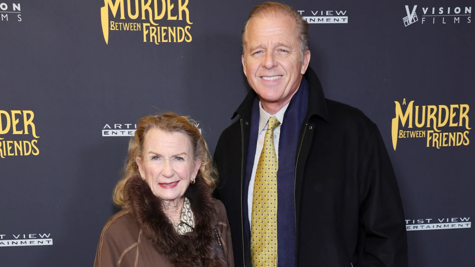 An older woman in a fur-trimmed coat and a man in a black jacket and yellow tie stand smiling in front of a "A Murder Between Friends" event backdrop.