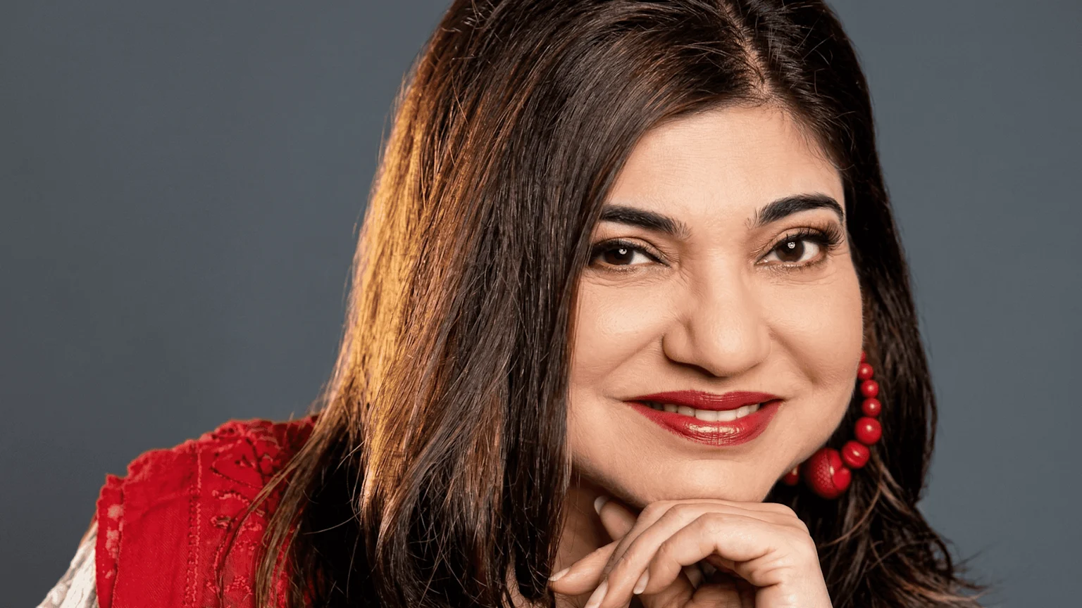 Alka Yagnik smiles cheerfully at the camera, wearing red lipstick and large red beaded earrings, with her hand resting under her chin against a grey background.
