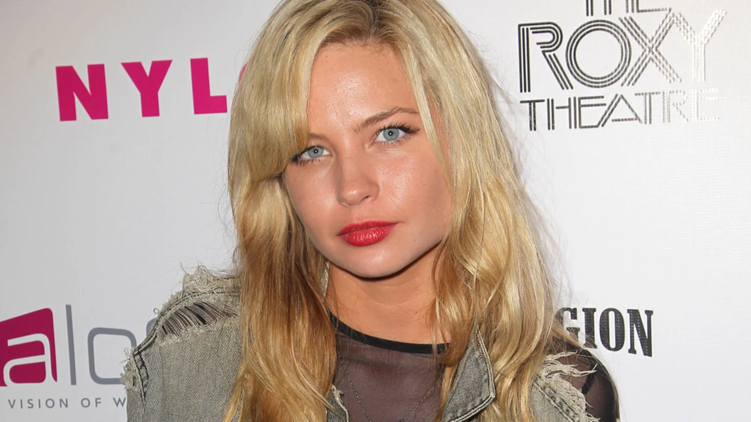 A blonde woman with blue eyes and red lipstick looks directly at the camera, wearing a distressed denim jacket, with "NYLON", "ROXY THEATRE", and "VISION OF W" logos visible in the background.