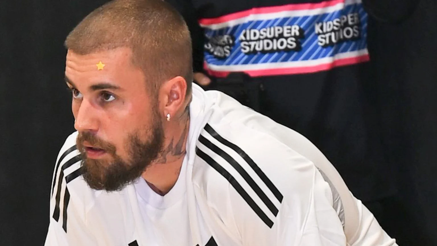 A man with a beard, short hair, neck tattoo, and an earring wears a yellow star sticker on his forehead and a white athletic shirt with black stripes, with a "KIDSUPER STUDIOS" banner in the background.