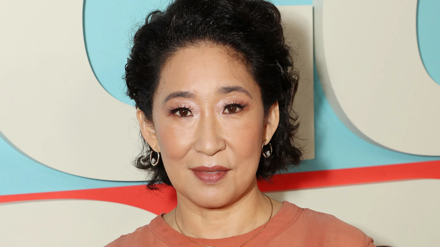 Sandra Oh, with dark curly hair pulled back, looks directly at the camera, wearing an earthy orange top and delicate hoop earrings against a light blue and off-white geometric background.