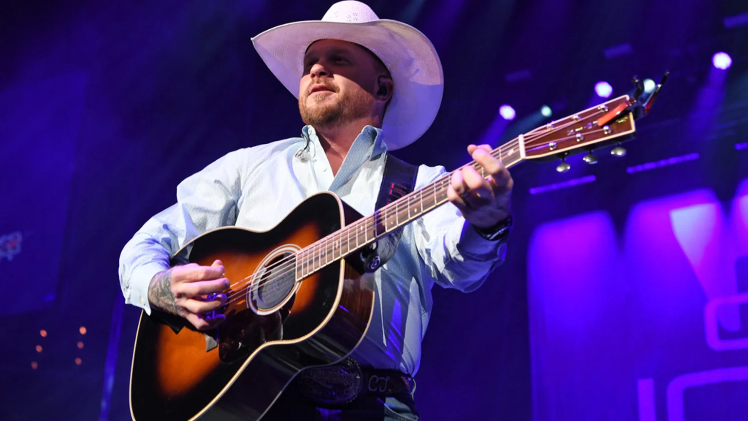 Cody Johnson, a man in a white cowboy hat and light blue shirt, plays an acoustic guitar on a stage bathed in purple light, with his tattooed left arm visible.