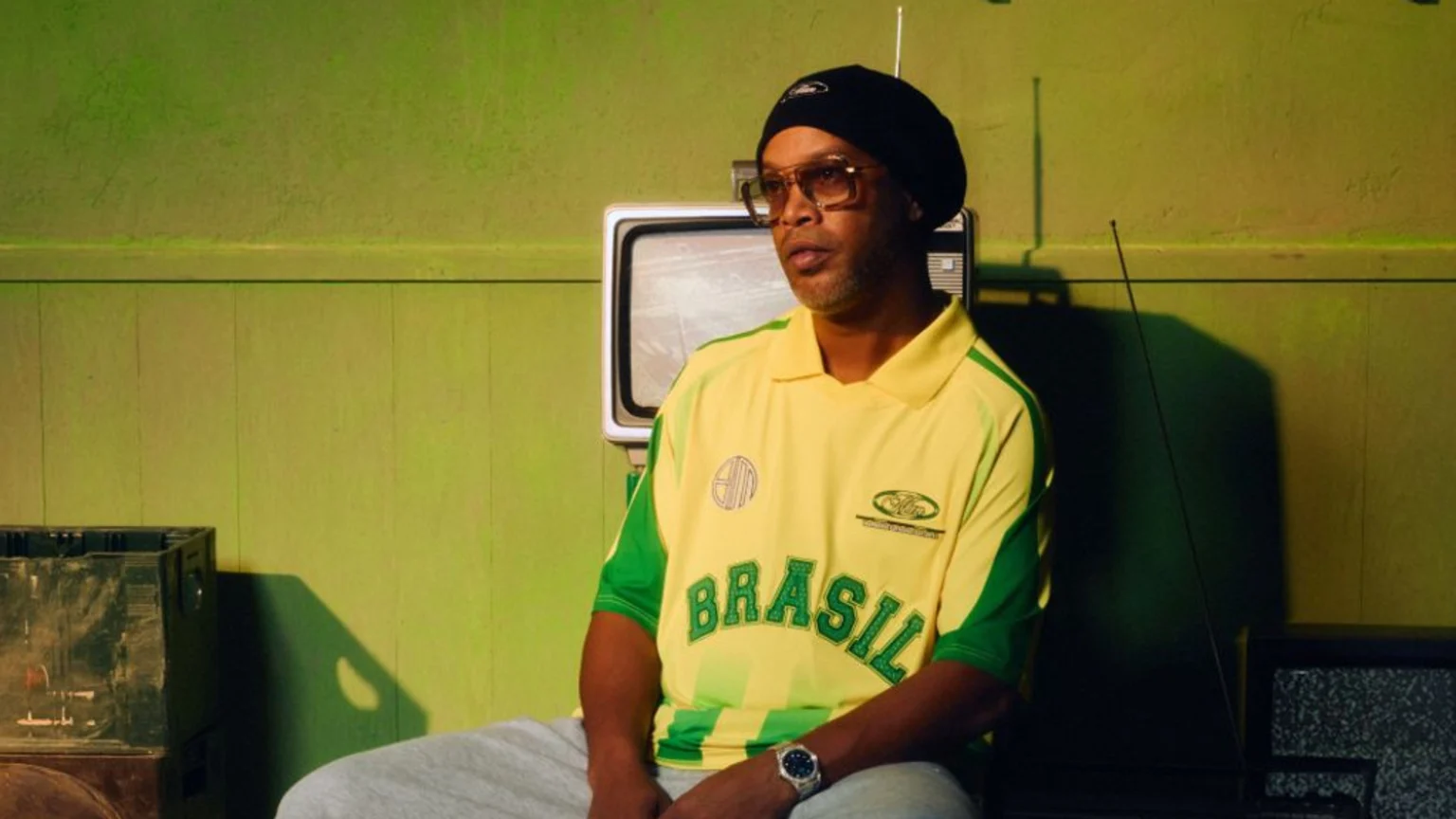 A man in a yellow and green "BRASIL" polo shirt, black beanie, and glasses sits, with a retro TV and a lime green wall behind him.