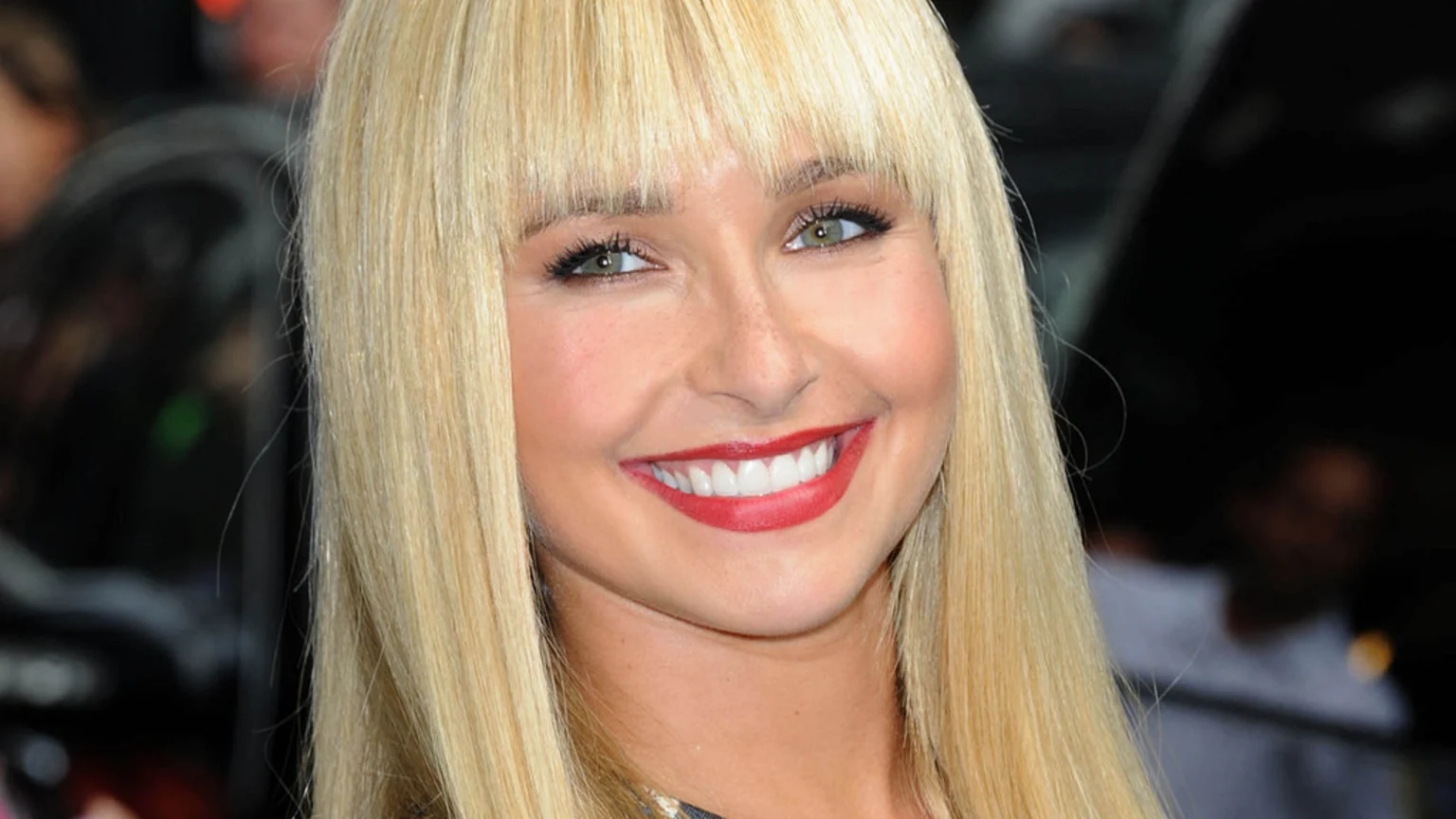 A close-up of a smiling woman with long blonde hair and bangs, green eyes, and red lipstick, showing some freckles.