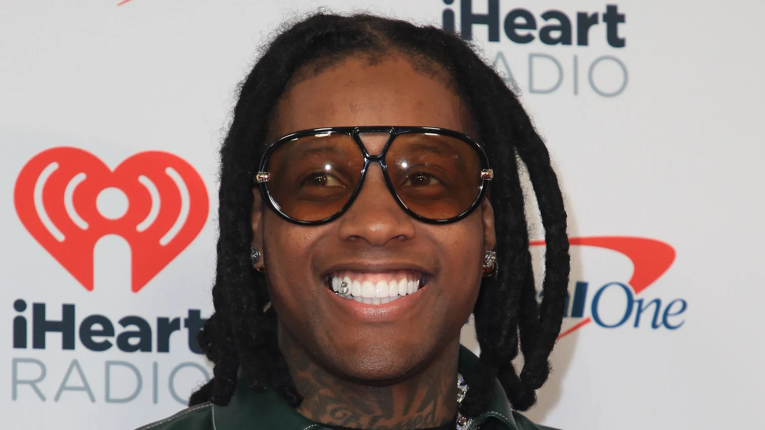 Close-up of a smiling person with dark dreadlocks and large brown sunglasses, showing white teeth with a single sparkling gem on a lower front tooth, against a white background with logos.