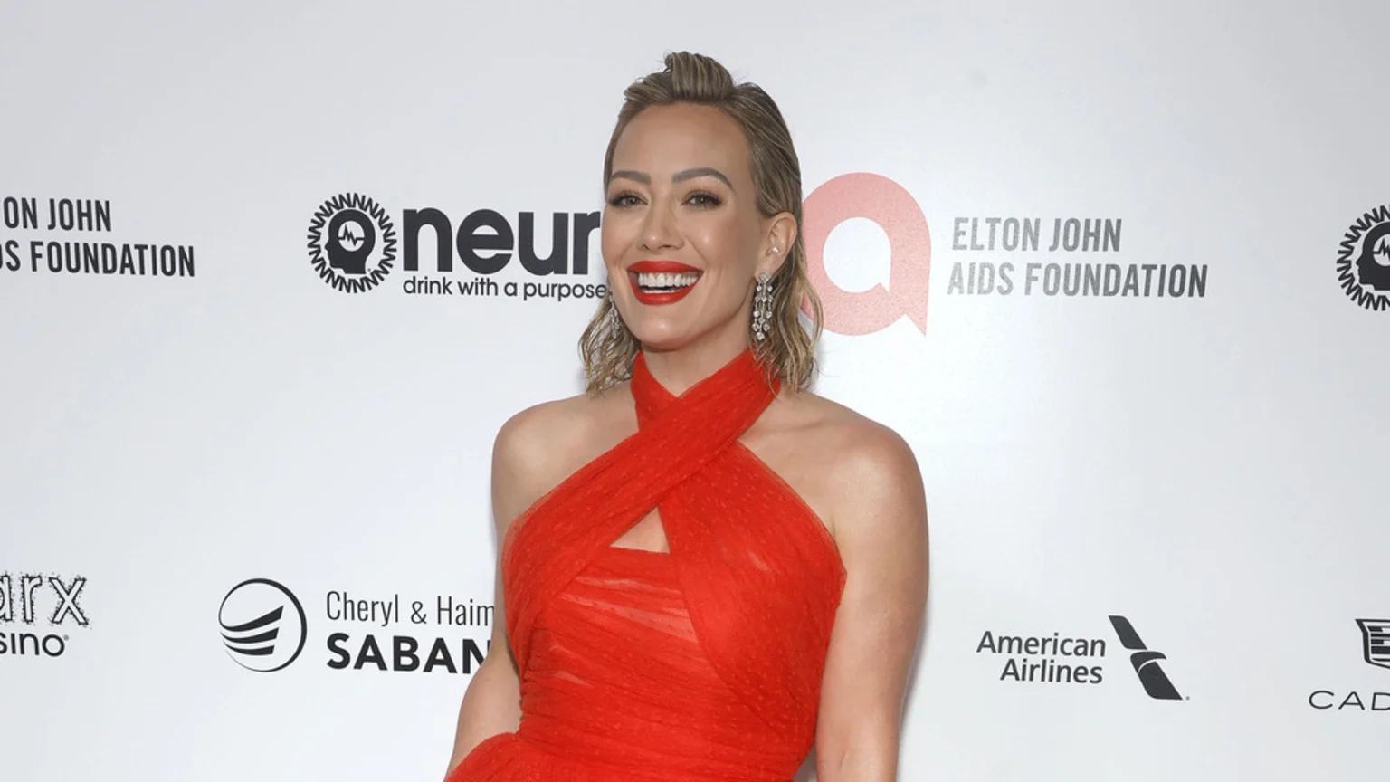 Hilary Duff smiles brightly in a red halter-neck gown, adorned with sparkling drop earrings and bold red lipstick, standing against a white backdrop with sponsor logos.