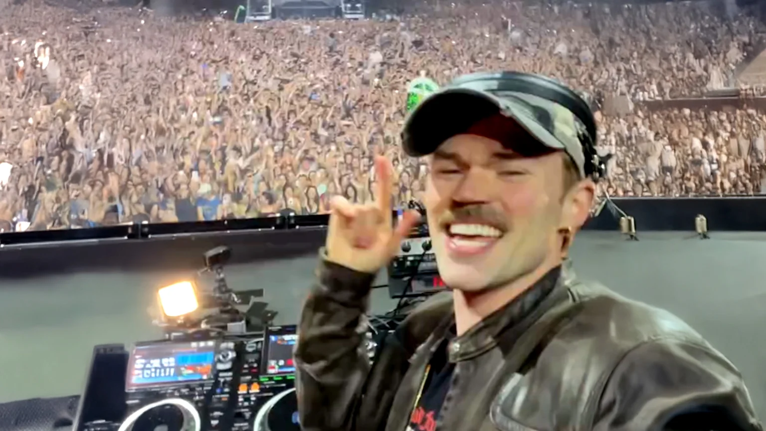 A smiling male DJ with a mustache and camo hat makes a rock-on gesture in front of his mixing equipment, with a vast, cheering crowd visible behind him at a night event.