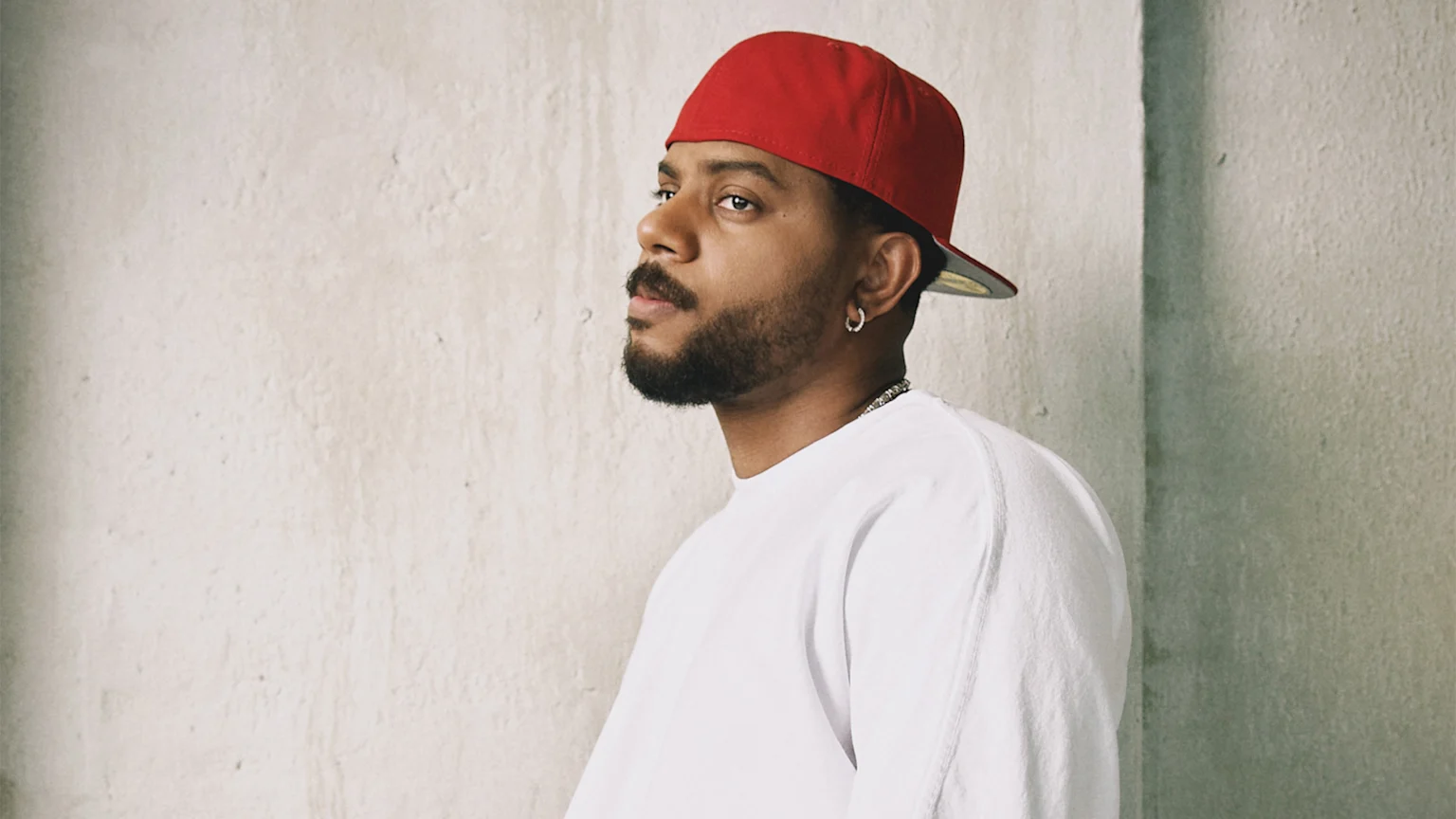A man with a beard and red baseball cap worn backward looks left, wearing a white t-shirt and silver chain, against a textured wall.