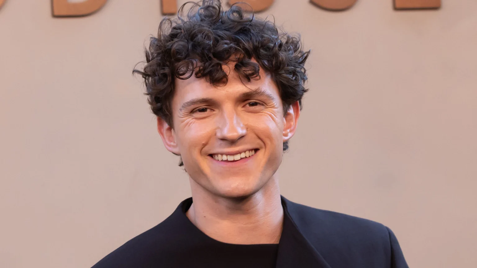 Tom Holland smiles broadly, showing his teeth, with curly dark hair and wearing a dark jacket against a beige background.
