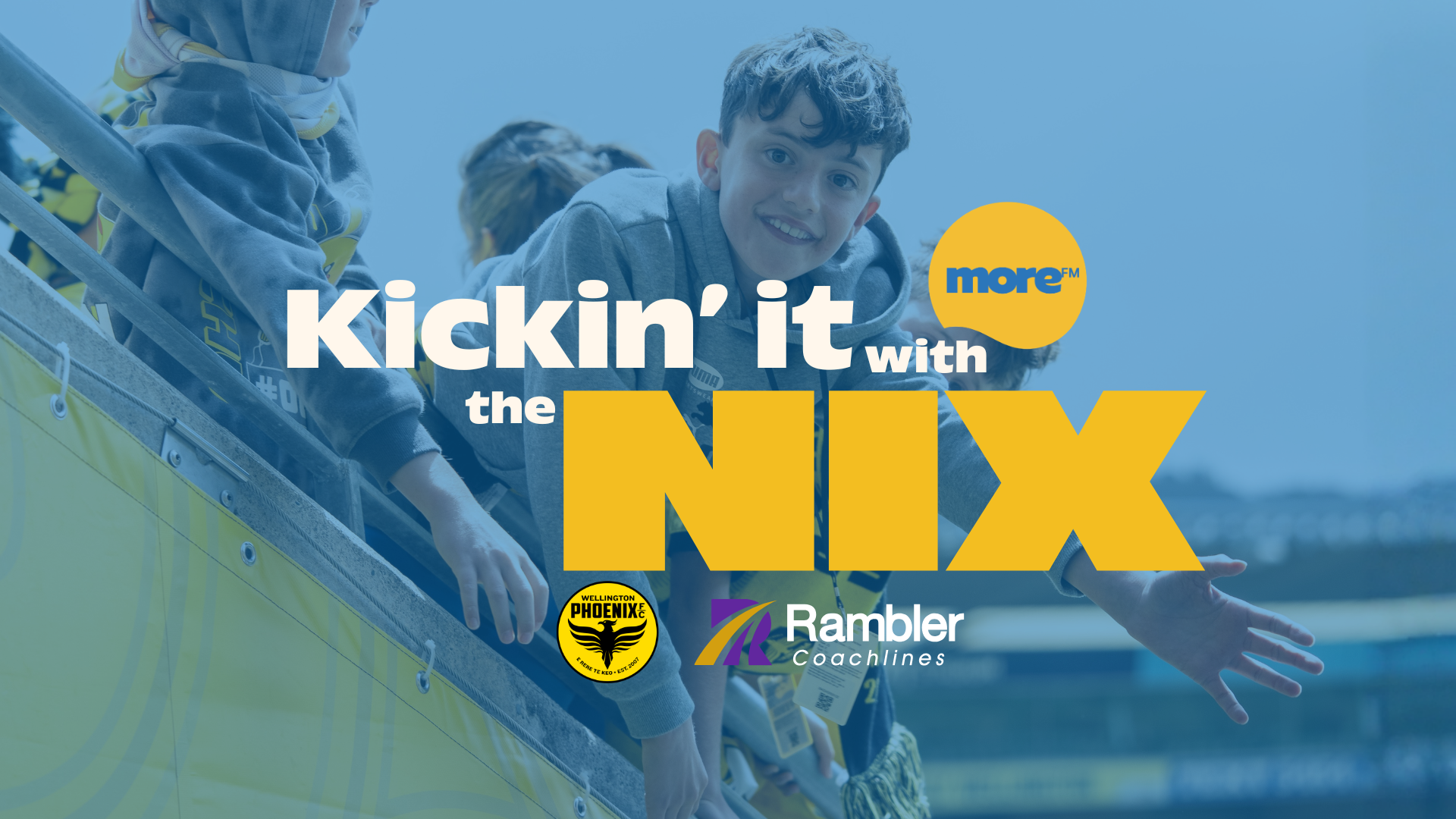 A smiling boy and other children lean over a stadium barrier. Overlaid text reads "Kickin' it with the NIX," with logos for More FM, Wellington Phoenix, and Rambler Coachlines against a blue background.