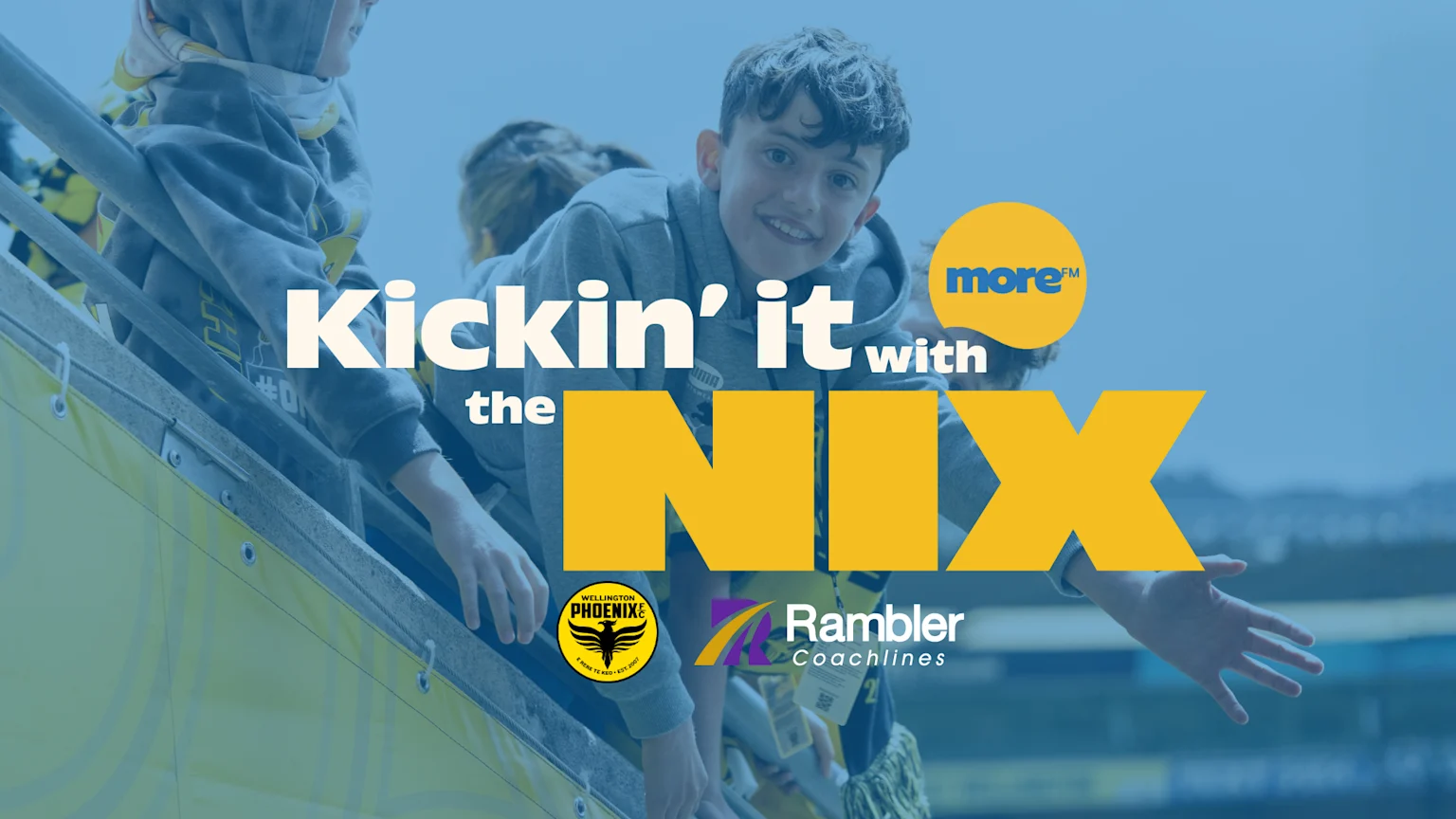 A smiling boy and other children lean over a stadium barrier. Overlaid text reads "Kickin' it with the NIX," with logos for More FM, Wellington Phoenix, and Rambler Coachlines against a blue background.