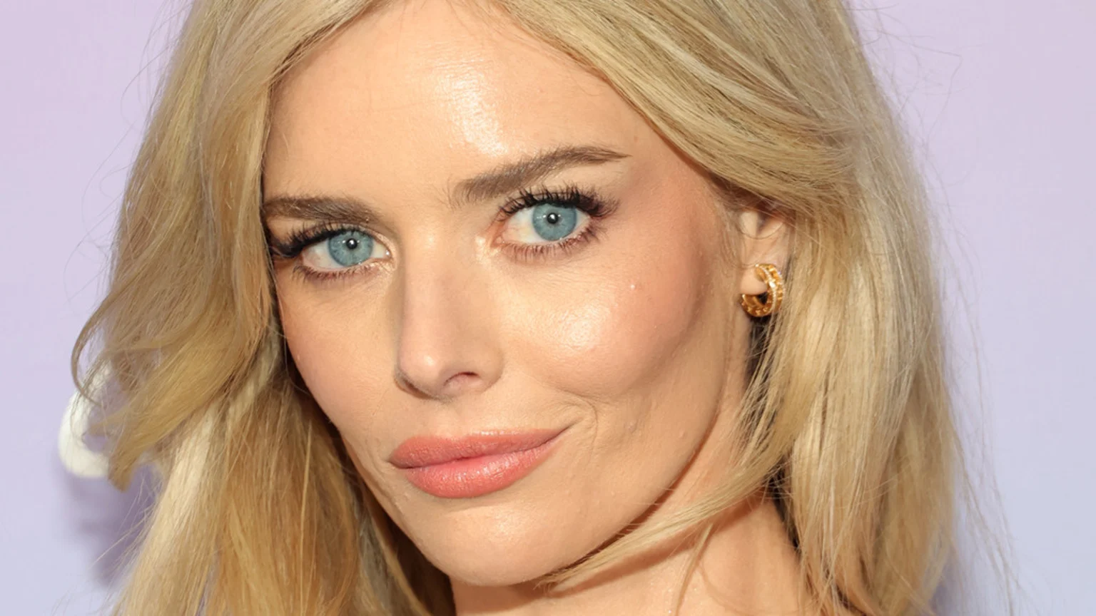 A close-up portrait of Samara Weaving, a blonde woman with blue eyes, light makeup, and a gold textured hoop earring, against a soft lavender background.