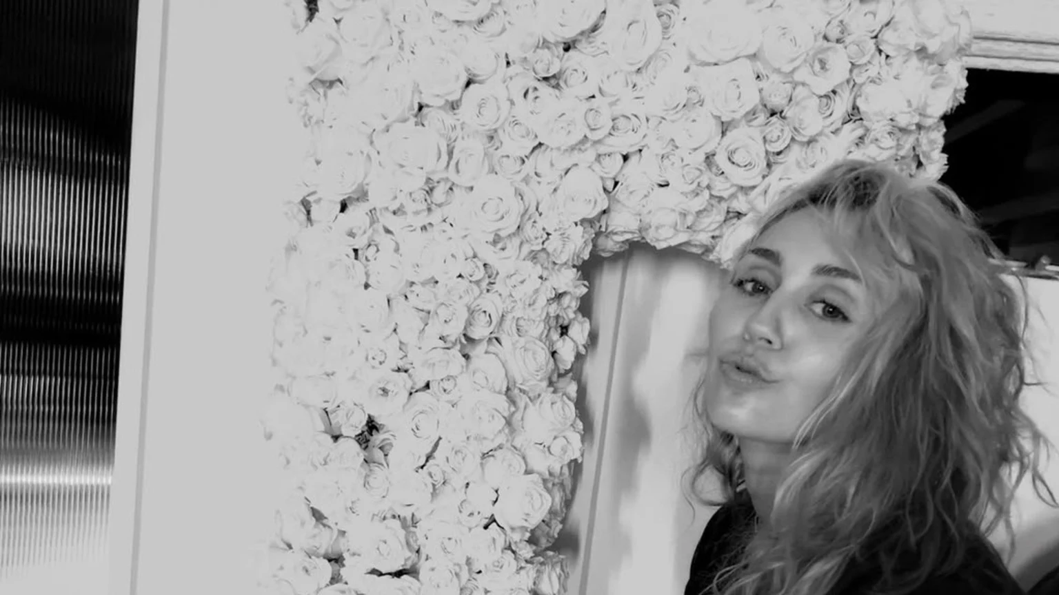 Black and white shot of a woman with wavy hair blowing a kiss, framed by a wall covered in white roses, with a dark textured panel on the left.