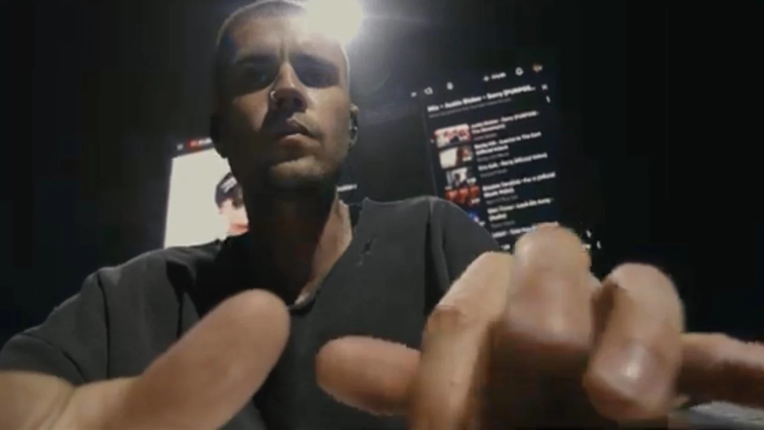 Justin Bieber looks directly at the camera, illuminated from above, with out-of-focus fingers close to the lens and a screen displaying a playlist with "Justin Bieber - Sorry" in the background.
