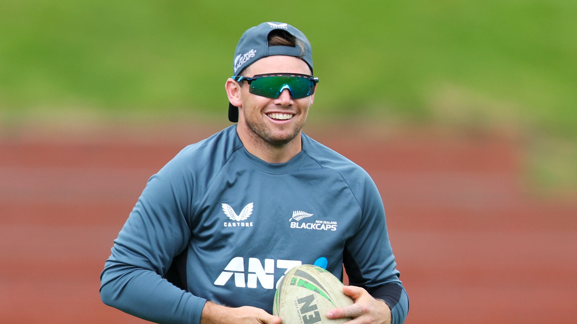 A smiling man in a dark teal long-sleeved athletic shirt with Blackcaps, Castore, and ANZ logos, reflective sunglasses, and a backward cap, holds a rugby ball against a blurred green field and red track.