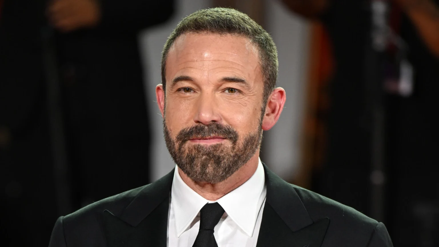 Close-up of actor Ben Affleck looking forward with a slight smile, wearing a black suit, white shirt, and black tie, with a short beard and mustache.