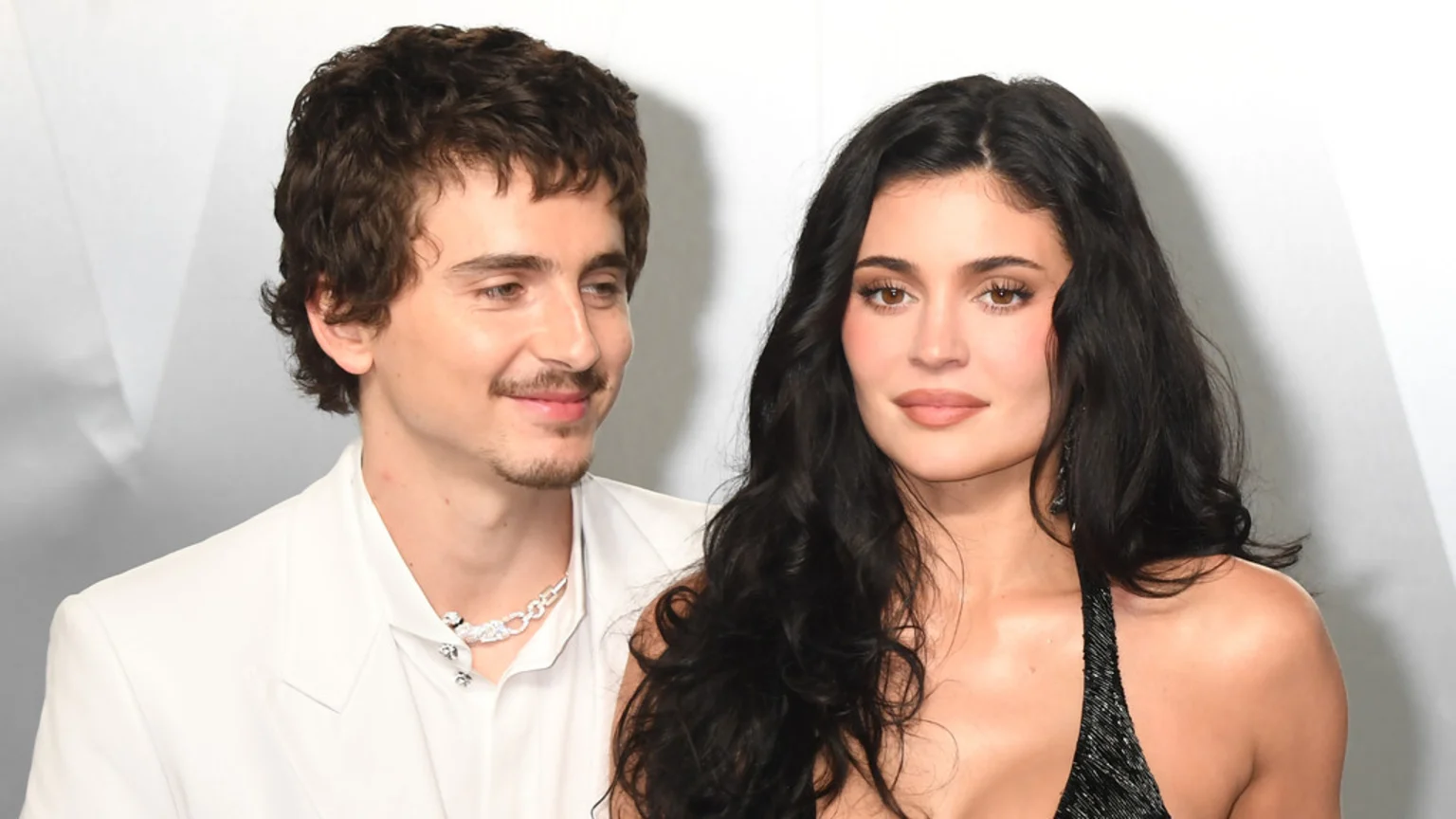 Kylie Jenner and Timothée Chalamet pose together, with Timothée in a white suit smiling subtly at Kylie, who looks forward with long dark hair and a dark sparkly top.