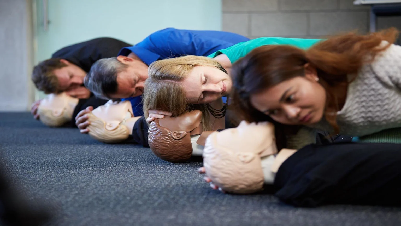 Several adults lie in a row on the floor, practicing CPR on individual training mannequins of varying complexions.