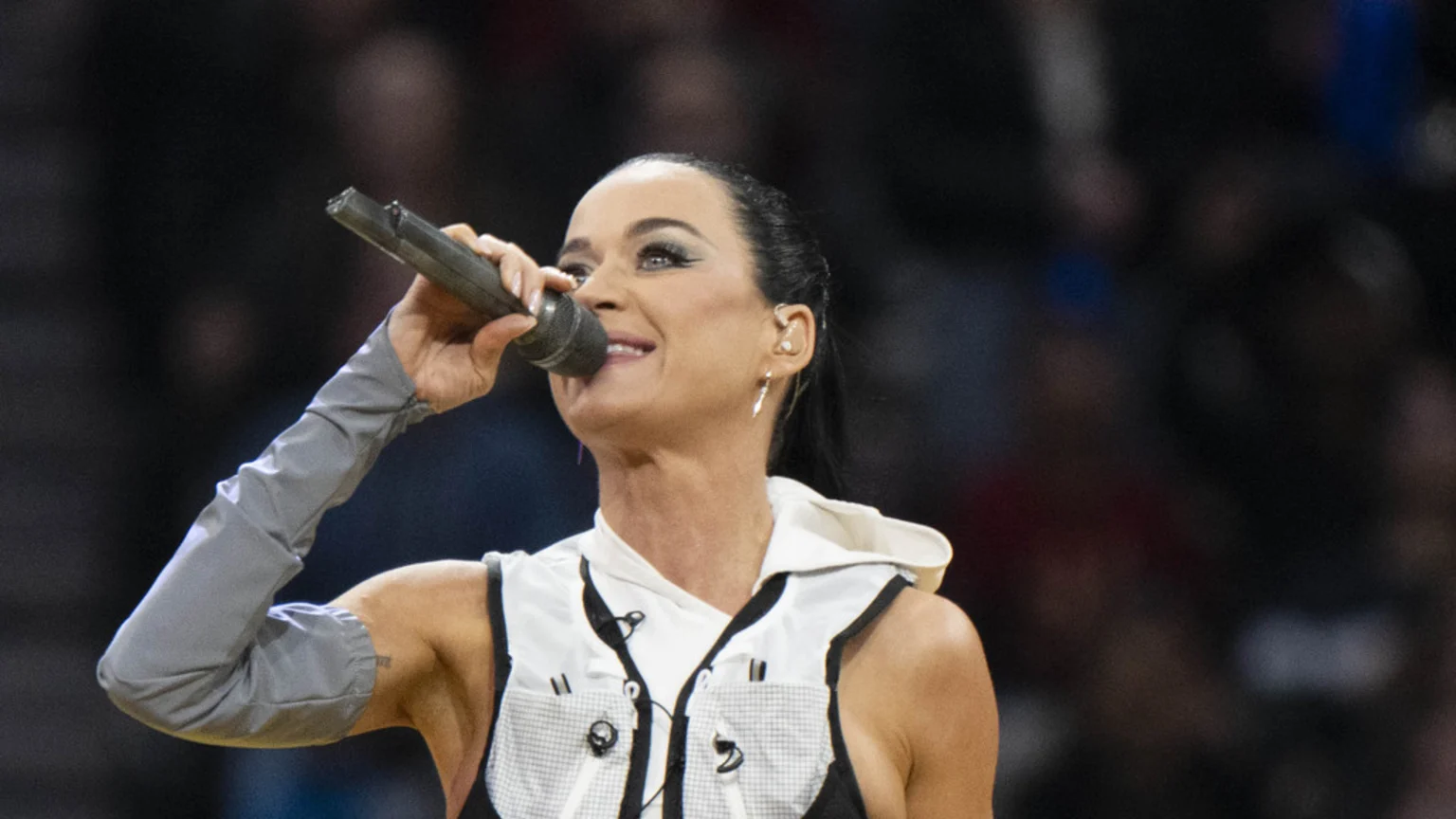 Katy Perry smiles while singing into a microphone, wearing a white hooded vest over a black top and a gray arm sleeve on her left arm, against a dark, blurred background.