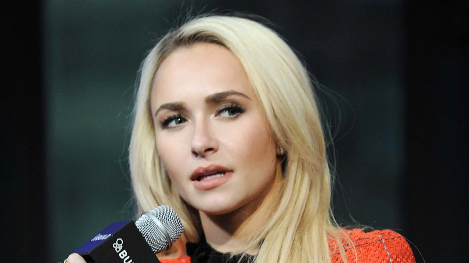 Hayden Panettiere, a blonde woman, speaks into a microphone, wearing an orange and black top against a dark background.