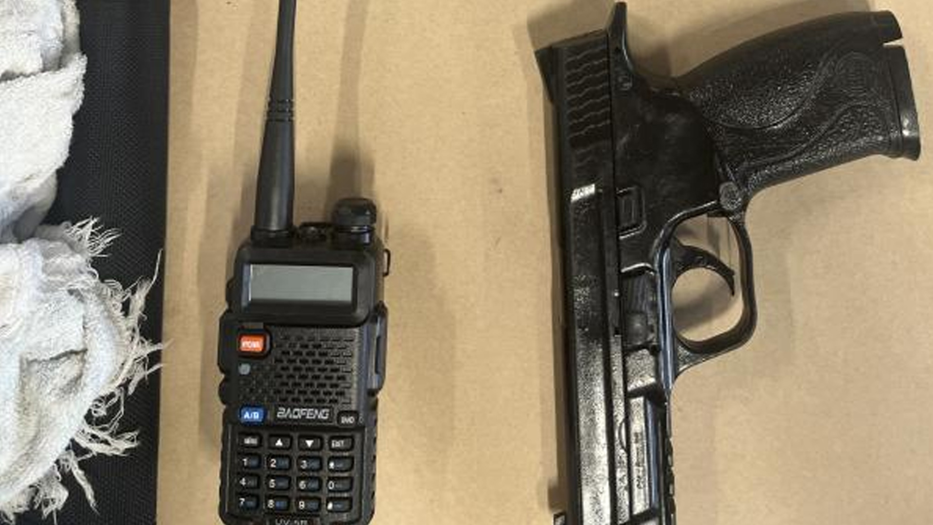 A black Baofeng UV-5R two-way radio, a black semi-automatic pistol, and white tattered cloth are laid out on a tan surface.