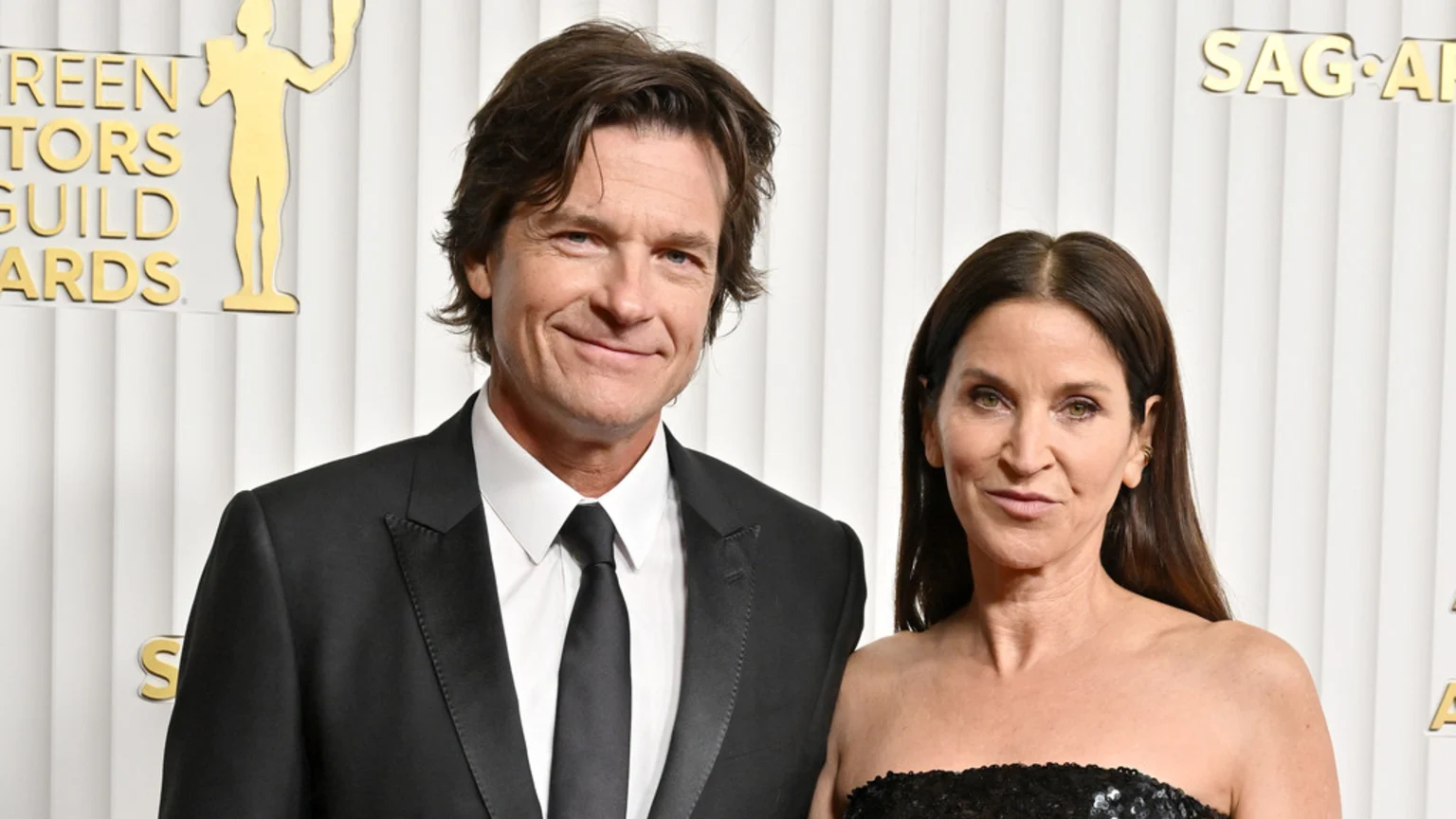 A formal portrait of actor Jason Bateman and his wife Amanda Anka posing at the Screen Actors Guild Awards. Jason wears a black suit and tie, Amanda a black strapless sequined top, against a white backdrop with gold SAG Awards lettering and statuette.
