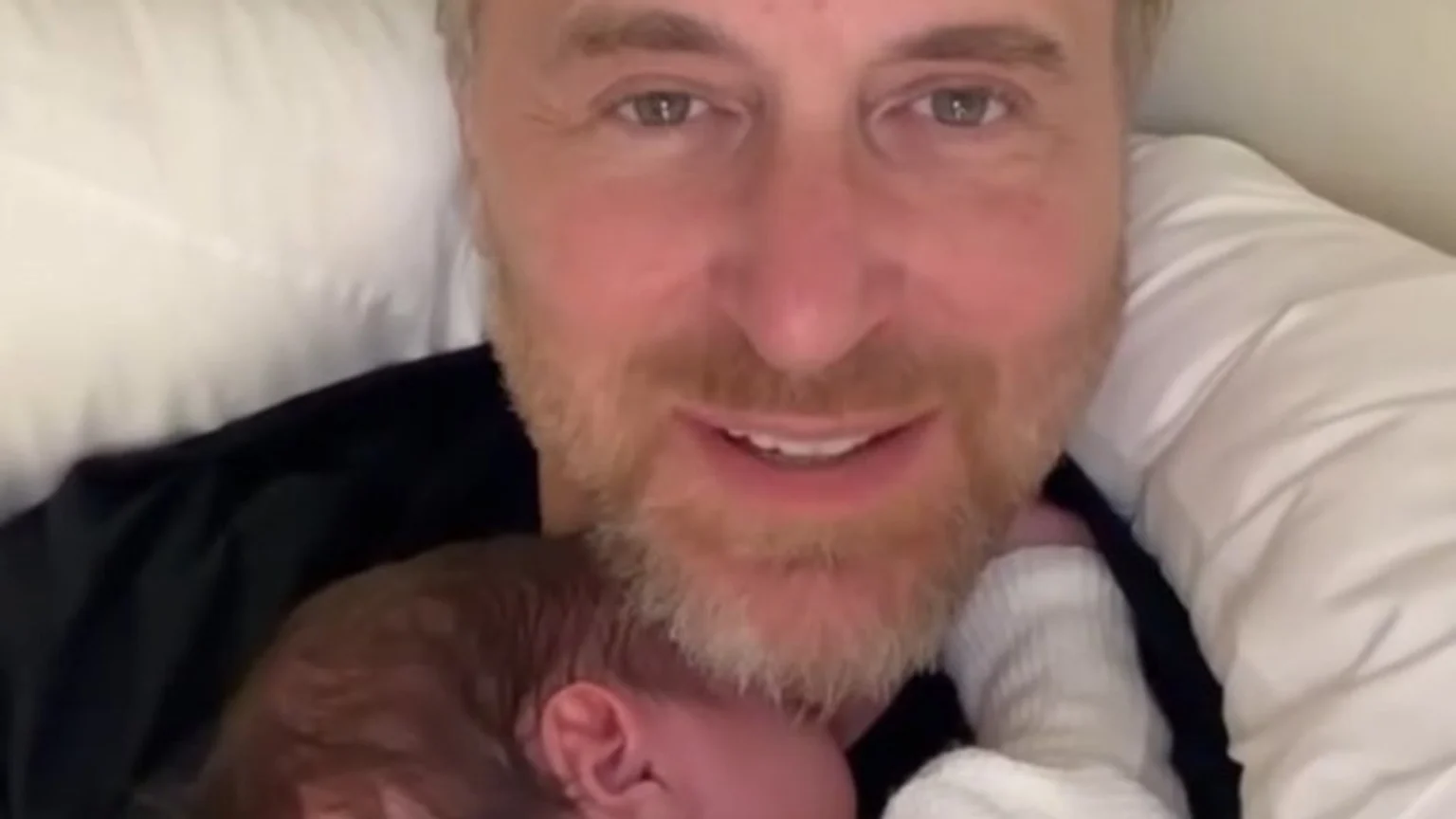 A bearded man smiles at the camera, holding a baby with reddish-brown hair nestled against his neck and chest.