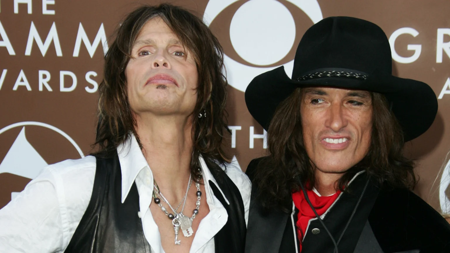 Aerosmith members Steven Tyler, with long hair and a white shirt, and Joe Perry, wearing a black cowboy hat and a red scarf, pose together in front of a Grammy Awards backdrop.