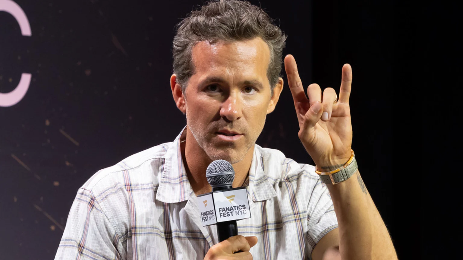 Actor Ryan Reynolds, with graying hair and stubble, speaks into a Fanatics Fest microphone while making a rock-on gesture with his right hand, wearing a light plaid shirt.