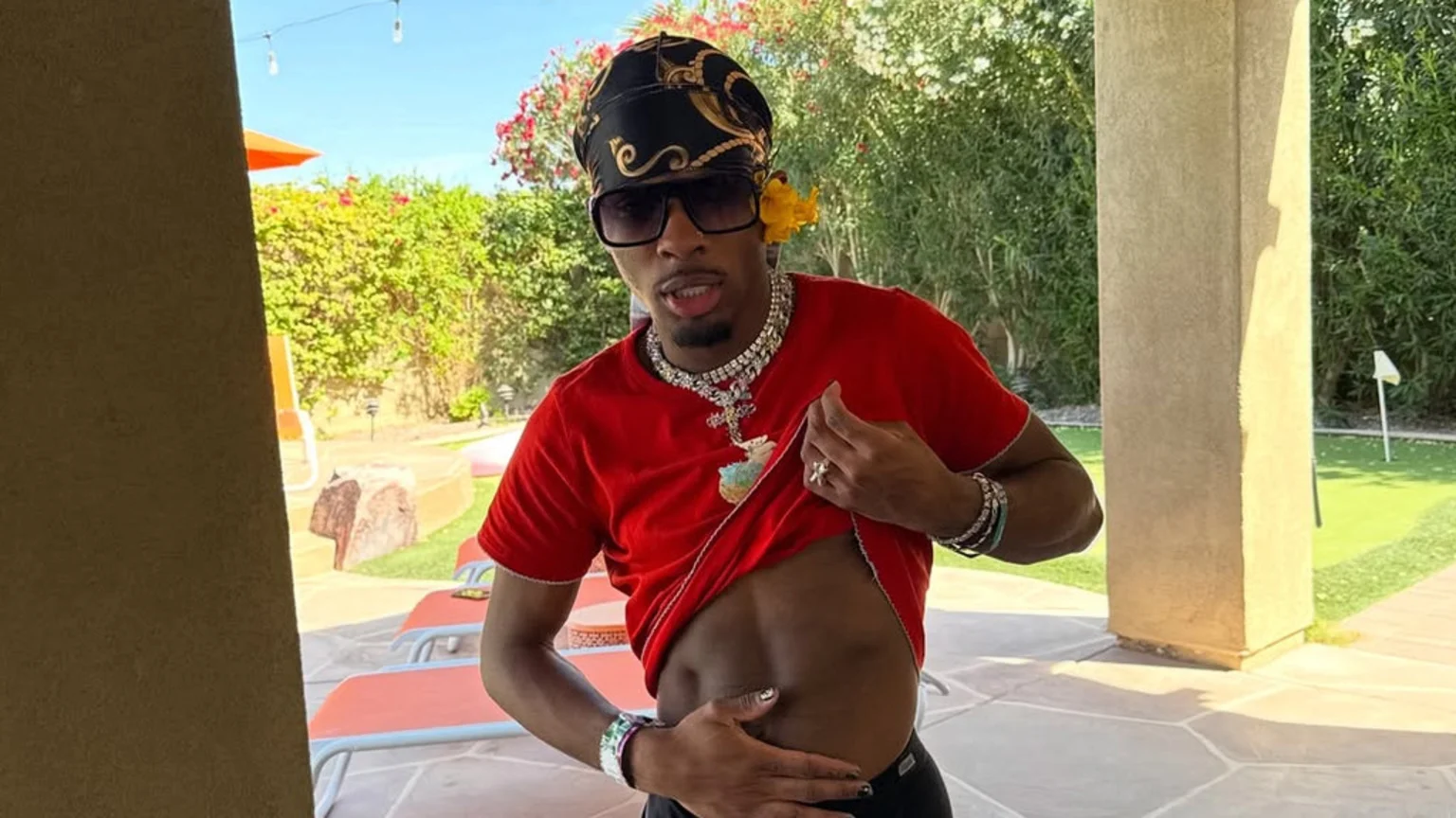 A man wearing a black headscarf, sunglasses, and multiple necklaces lifts his red t-shirt to reveal his abdomen while standing on a patio with greenery and a putting green in the background.