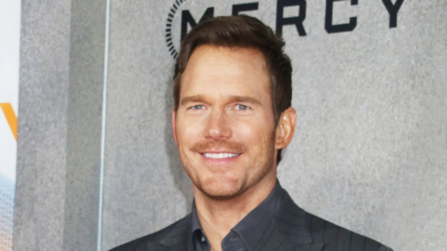 Chris Pratt smiles and looks at the camera, wearing a dark suit jacket and shirt, with a blurred "MERCY" sign visible in the background.