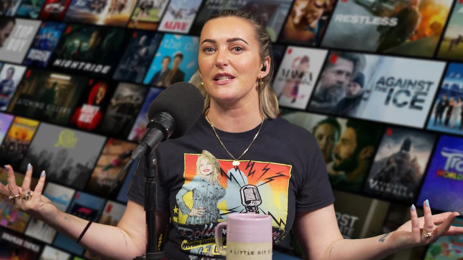 A woman with light brown hair speaks into a microphone, wearing a Dolly Parton t-shirt, with a blurred background showing a grid of Netflix movie and TV show titles.