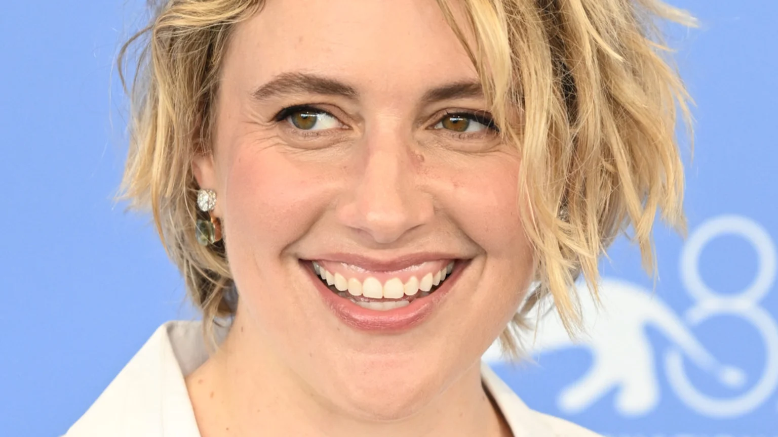 Close-up of Greta Gerwig smiling broadly, with short, messy blonde hair, brown eyes, and earrings, against a light blue background.
